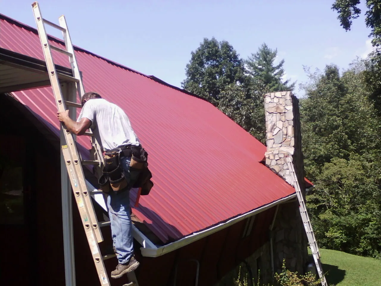 Roofer on a ladder beside a red metal roof and stone chimney — Soffit & Fascia Repair by Roofing Brothers in Roebling